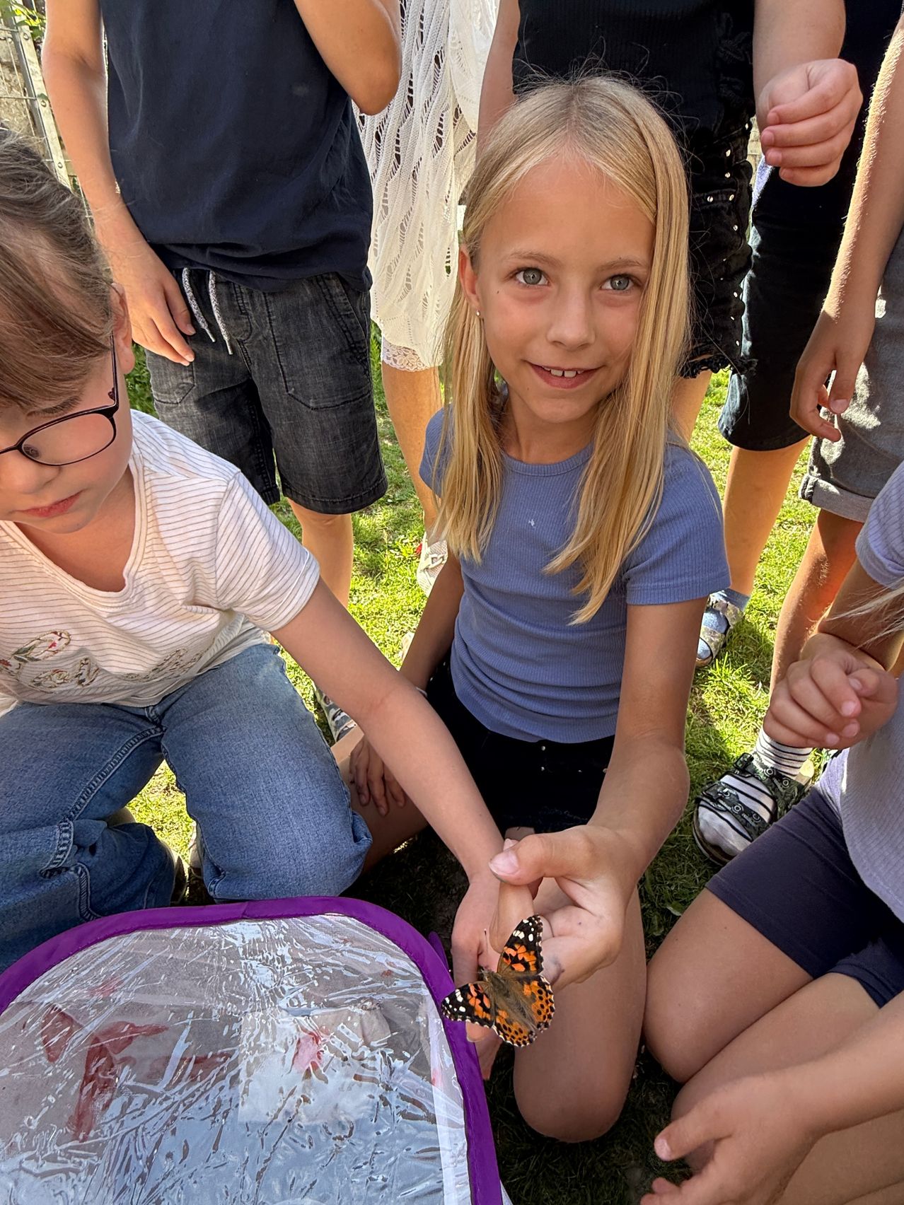Zwei Kinder beobachten einen Schmetterling mit einer transparenten Hülle zwischen ihnen auf einem Grasfeld. Ein Mädchen in einem blauen Shirt hält den Schmetterling.