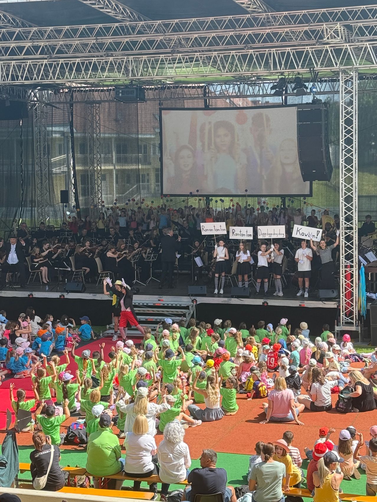 Eine große Menschenmenge in grünen Shirts und Hüten versammelt sich auf einer Bühne. Vor ihnen steht ein großes Orchester mit Musikern. Im Hintergrund befindet sich eine große Leinwand. Einige Leute halten Schilder mit Fragen hoch.