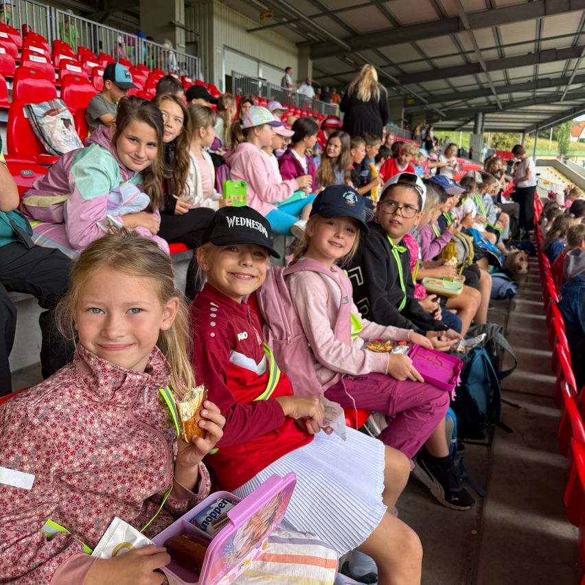 Eine Gruppe junger Mädchen mit verschiedenen Snacks und Schulsachen sitzt in einem Stadion. Einige tragen Jacken und Mützen. Dahinter sitzen weitere Kinder in Reihen, wobei gelegentlich Erwachsene zu sehen sind.
