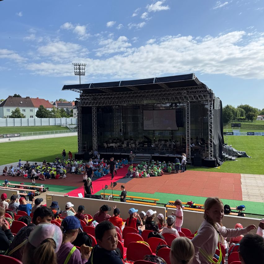 Eine große Menschenmenge aus Kindern und Erwachsenen sitzt auf roten Stühlen und beobachtet eine Aufführung auf einer Bühne. Die Bühne hat eine große Leinwand, Lautsprecher und einen roten Teppich. Hinter der Bühne befindet sich ein Rasenfeld und ein Gebäude.