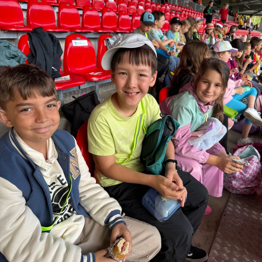 Drei Kinder sitzen zusammen in einem Stadion. Zwei Jungen und ein Mädchen, sie haben Snacks und tragen Jacken. Leere rote Sitze sind hinter ihnen.