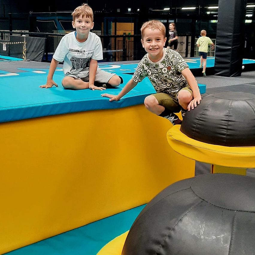 Zwei junge Jungen spielen auf einem Trampolin in einem bunten Indoor-Spielplatz, ein Junge sitzt und der andere lehnt sich an einen gelben Ball.