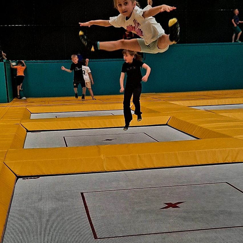 Ein Mädchen springt in der Luft auf einem Trampolin, während andere zusehen. Der Boden ist gelb und grau mit einem roten X.