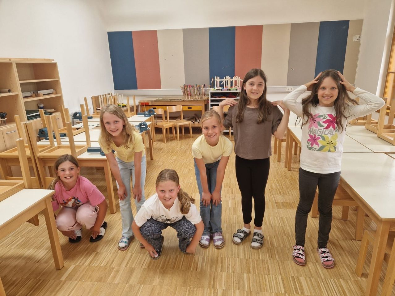 Fünf Mädchen in einem Klassenzimmer mit bunten Wänden posieren für ein Foto, lächeln und knien.