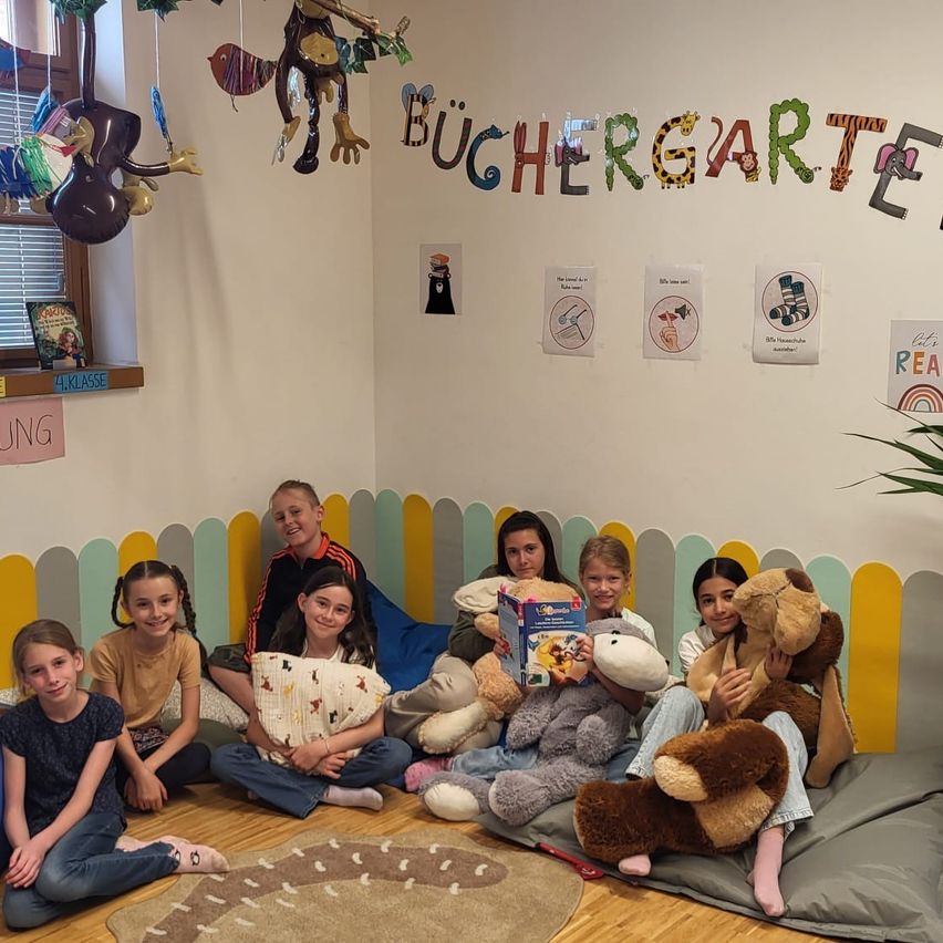 Eine Gruppe von Kindern sitzt in einer gemütlichen Leseecke auf dem Boden mit Plüschtieren und lächelt. Der Raum hat bunte Wanddekorationen und Schilder an der Wand. Ein Buch ist auf einem nahen Regal sichtbar.