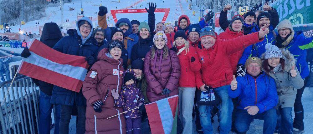 Eine Gruppe von Menschen steht im Schnee und posiert für ein Foto. Sie tragen Winterkleidung, einige mit Mützen und Handschuhen. Sie halten Flaggen und eine Handtasche. Einige von ihnen lächeln und einer hebt die Hand. Hinter ihnen steht ein verschneiter Berg mit Bäumen.