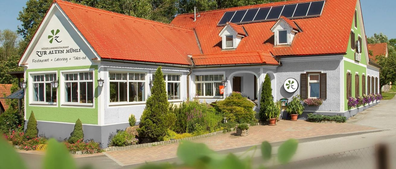 Ein weißes Gebäude mit rotem Dach und Solarpanelen, umgeben von einem Garten mit verschiedenen Pflanzen und Blumen. Das Gebäude hat ein Schild, das 'Mühle' und 'Take-out' besagt.