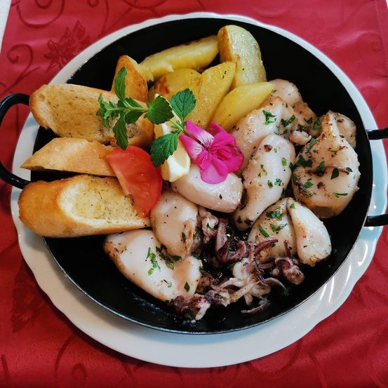 Eine schwarze Pfanne mit einem Meeresfrüchtegericht, bestehend aus Jakobsmuscheln, Tintenfisch und Kartoffeln, garniert mit Kräutern und einer rosa Blume, serviert auf einem weißen Teller mit Brot auf einem roten Tischtuch.