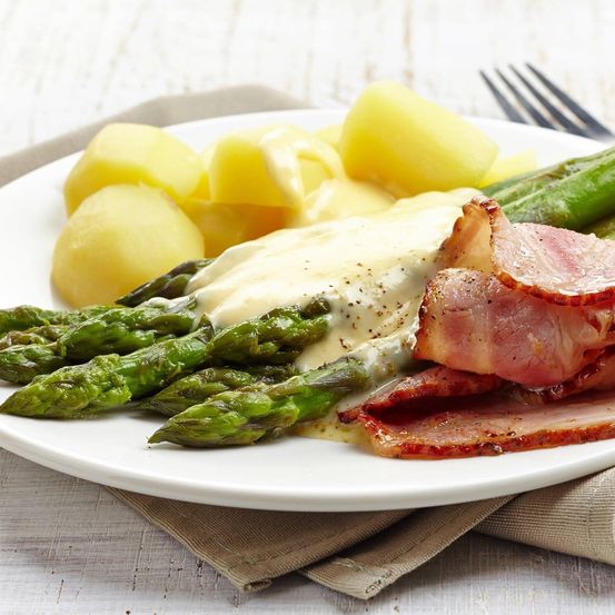 Ein Teller mit Spargel, Kartoffeln und Speck, überzogen mit einer cremigen Sauce. Das Gericht ist mit einer Zitronenscheibe garniert.