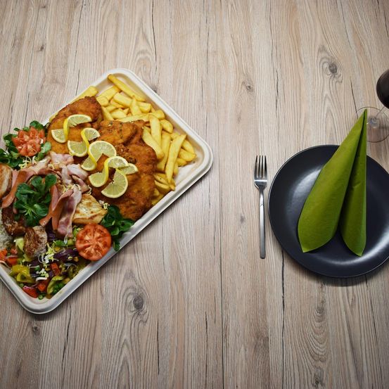 Ein weißer rechteckiger Teller mit verschiedenen Speisen wie gebratenem Hähnchen, Pommes Frites und Gemüse steht auf einem Holztisch. Daneben befindet sich ein schwarzer Teller mit einer grünen Serviette.