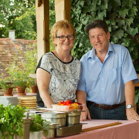 Ein Mann und eine Frau stehen hinter einem Tisch voller verschiedener Gemüsesorten und einer Schüssel bunter Früchte und lächeln für ein Foto in einer Gartenumgebung.