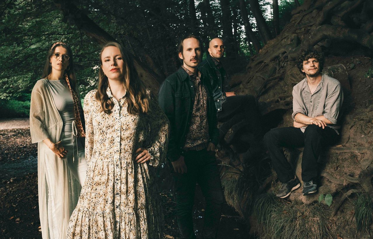 Eine Band aus vier Personen, darunter eine Frau und drei Männer, steht in einem Wald mit Bäumen und Felsen. Sie posieren für ein Foto.