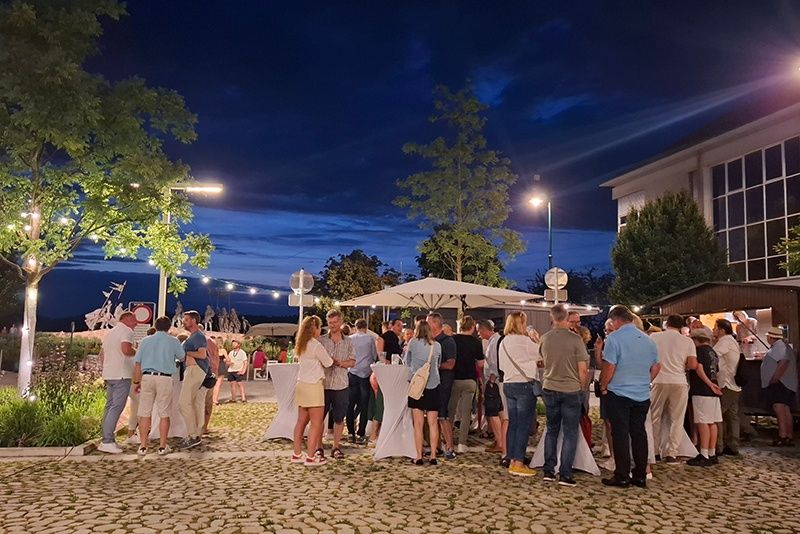 Eine Gruppe von Menschen versammelt sich bei einer Outdoor-Veranstaltung bei Nacht, unter Schirmen und in der Nähe eines Tisches, umgeben von Bäumen und Gebäuden.
