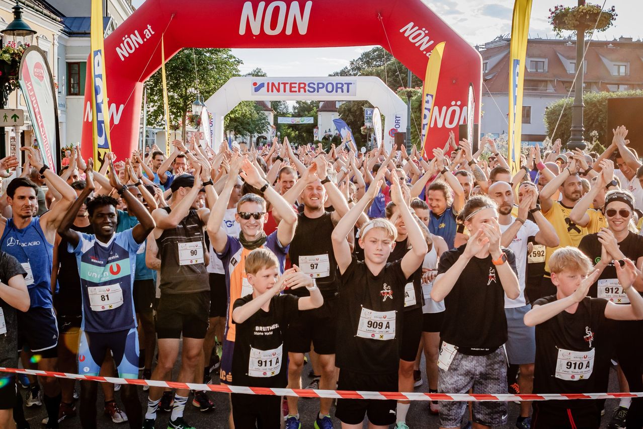 Eine Gruppe von Läufern versammelt sich an der Startlinie eines Rennens, jubelt und hebt die Hände. Sie tragen nummerierte Startnummern und sportliche Kleidung. Der Bogen über ihnen trägt die Aufschrift 'NON' und 'INTERSPORT.' Bäume und Gebäude sind im Hintergrund zu sehen.