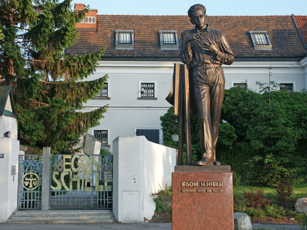 A bronze statue of Egon Schiele stands in front of a building with a tiled roof. The statue is on a pedestal with graffiti on the gate.