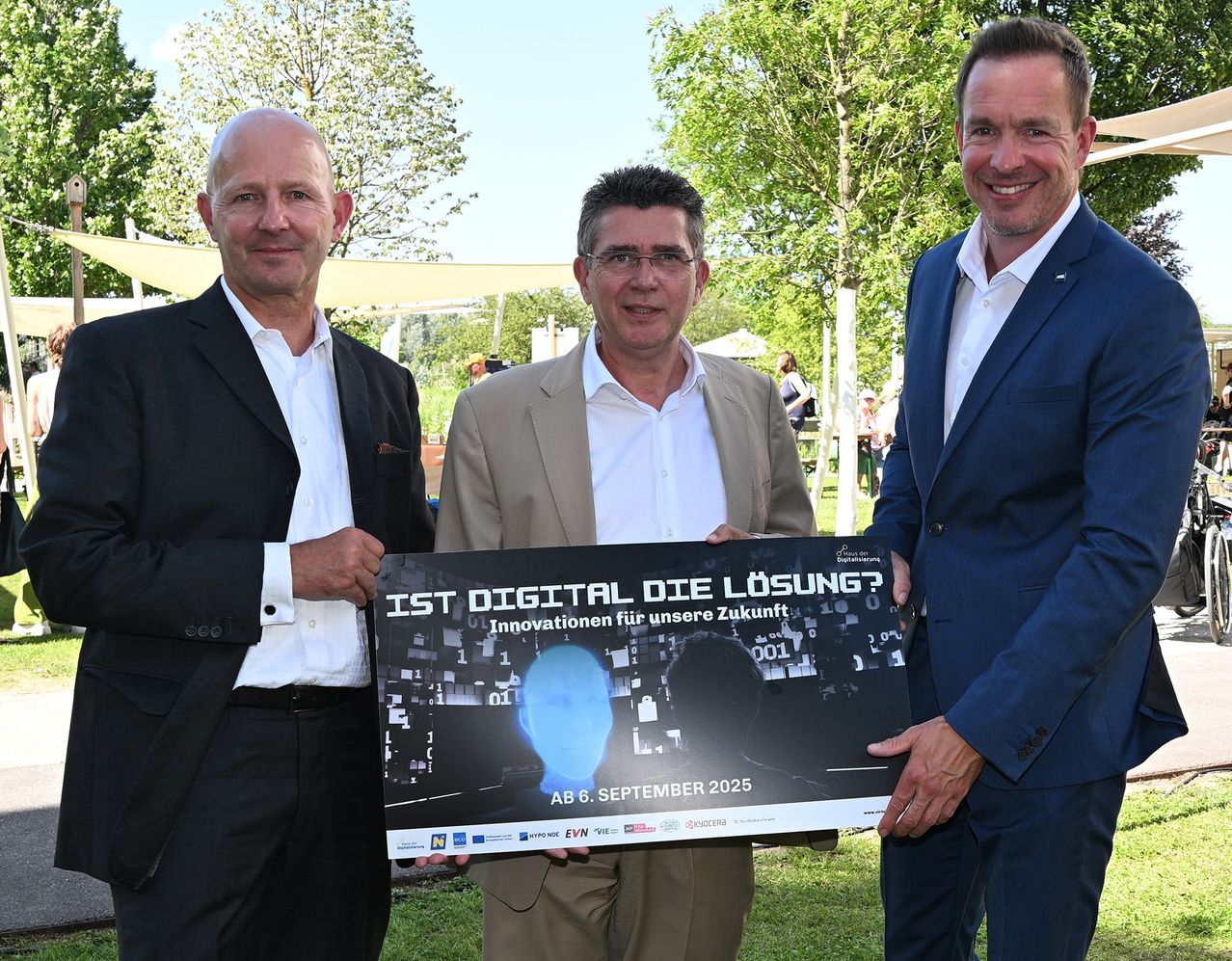 Three men in formal attire are standing in a park, holding a banner that reads 'IST DIGITAL DIE LÖSUNG?' The banner features a blue brain icon and mentions a date of September 6, 2025.