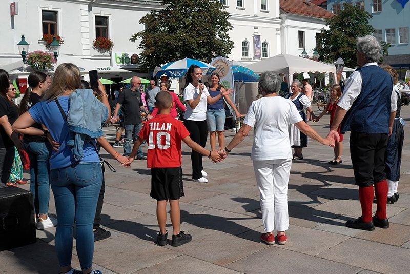 A group of people, including adults and children, are holding hands and dancing in an outdoor area. A woman is holding a microphone.