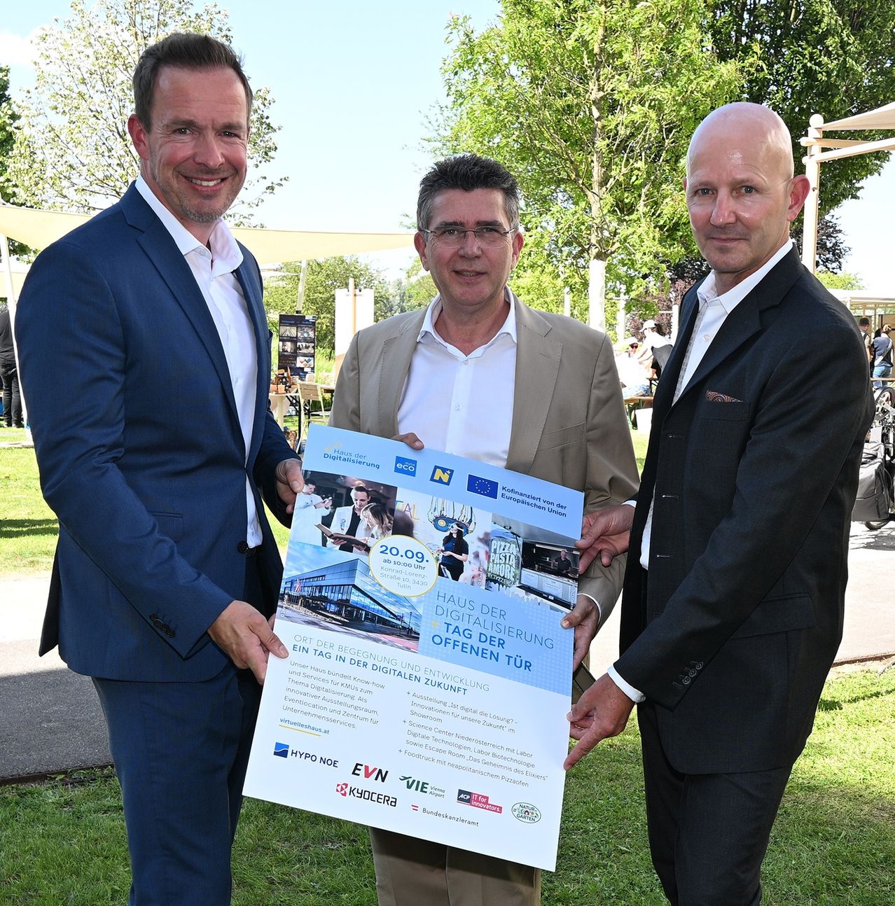 Three men stand in a park, one holding a poster that reads 'House of Digitalization'. The man on the left wears a blue suit, while the others wear white and black suits. They are surrounded by trees and a white tent.