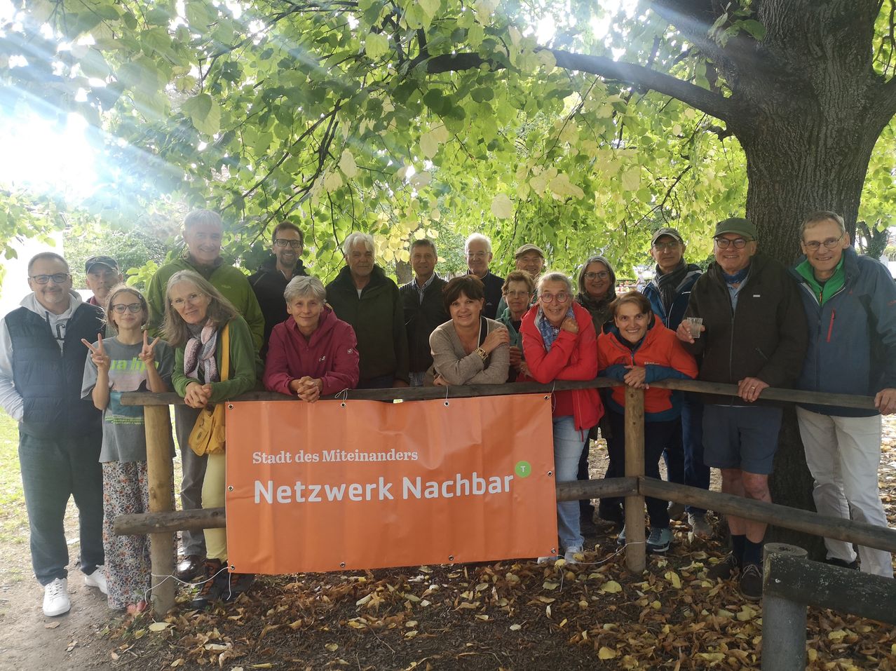 Eine Gruppe von Menschen posiert unter einem Baum mit einem orangefarbenen Banner, das 'Netzwerk Nachbar' und 'Stadt des Miteinanders' zeigt. Einige Leute lächeln und machen Gesten.