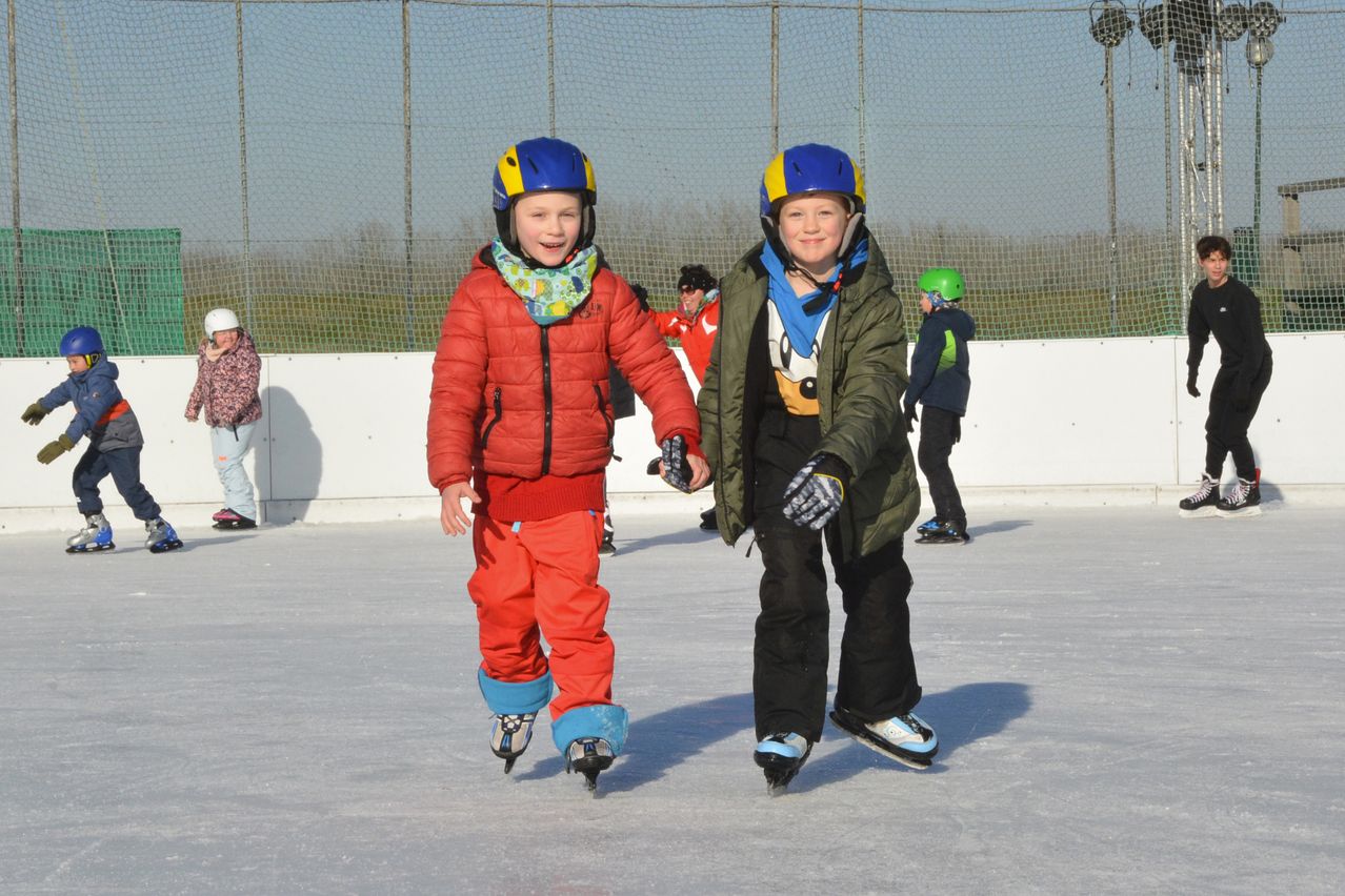 Zwei Kinder in Winterkleidung und Schlittschuhen rutschen gemeinsam auf einer Eisbahn. Hinter ihnen rutschen weitere Kinder und andere stehen in der Nähe der Barriere.