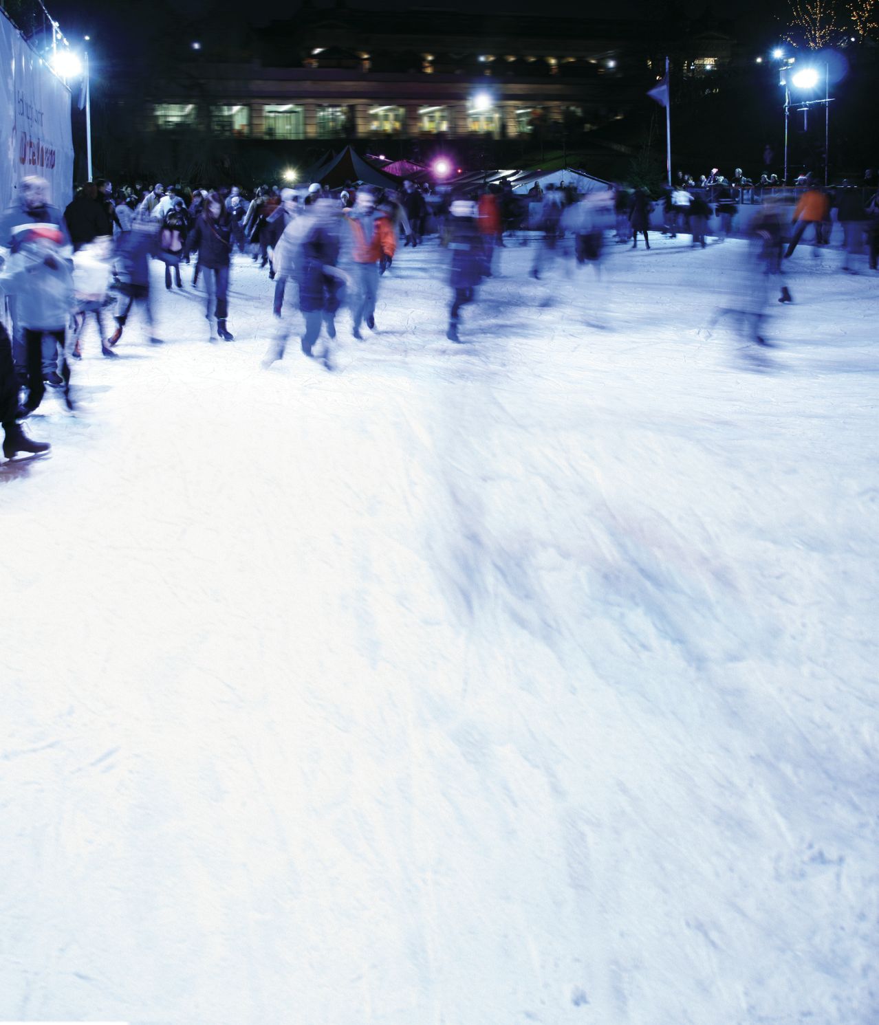 Viele Menschen skaten bei Nacht auf einer verschneiten Eisbahn, beleuchtet von Straßenlaternen. Gebäude und Zelte sind im Hintergrund sichtbar.