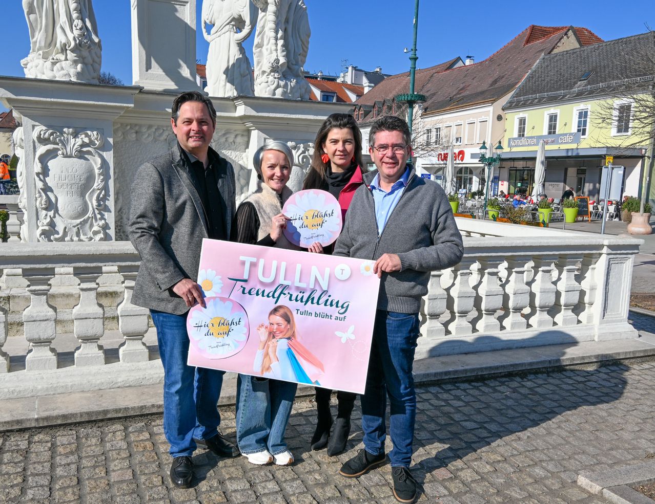 Vier Personen posieren vor einer Brücke mit einem rosa Schild, das Tulln Trendfruhling sagt, sie alle halten rosa Schilder. Hinter ihnen steht eine Statue, ein Gebäude und ein Café.