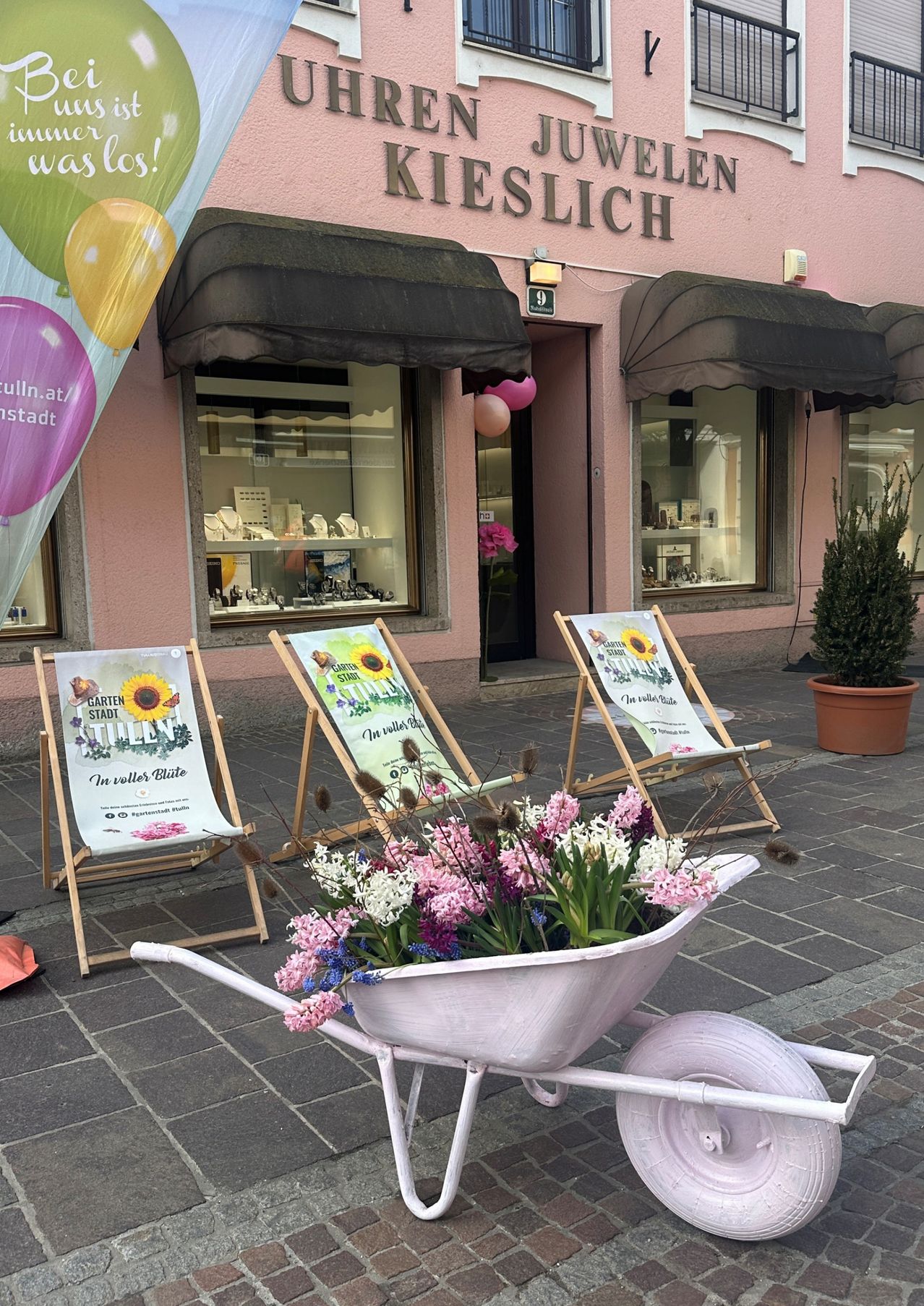 Das Schaufenster eines Ladens namens Kieslich ist mit Blumen, Luftballons und dekorativen Stühlen geschmückt. Ein rosafarbener Schubkarren voller Blumen steht davor.
