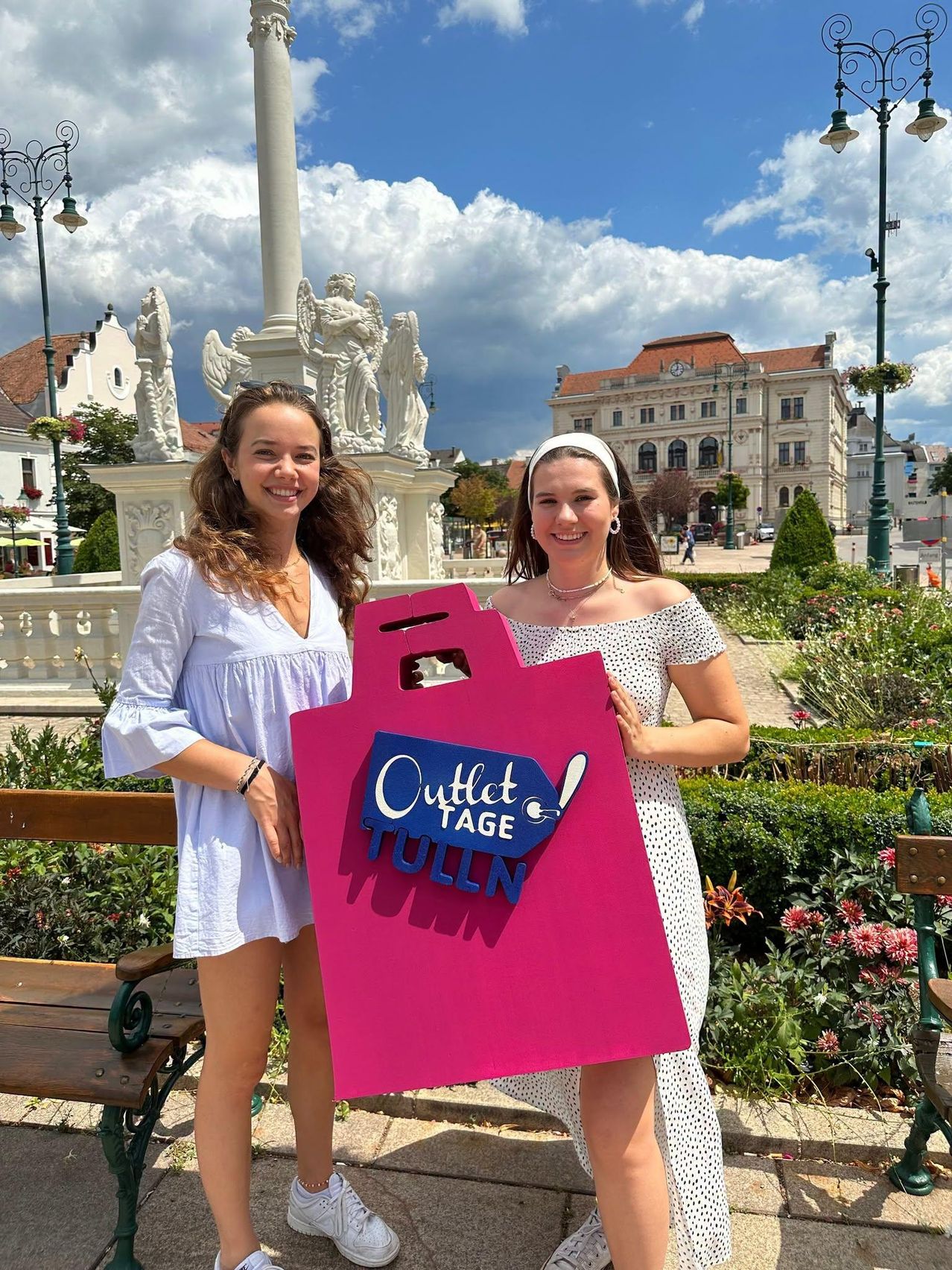 Zwei Frauen stehen in einem Park und halten eine rosa Tasche mit den Worten 'Outlet Tage Tulln'. Sie lächeln und scheinen für ein Foto zu posieren. Dahinter steht eine Statue und ein Gebäude mit einem roten Dach.