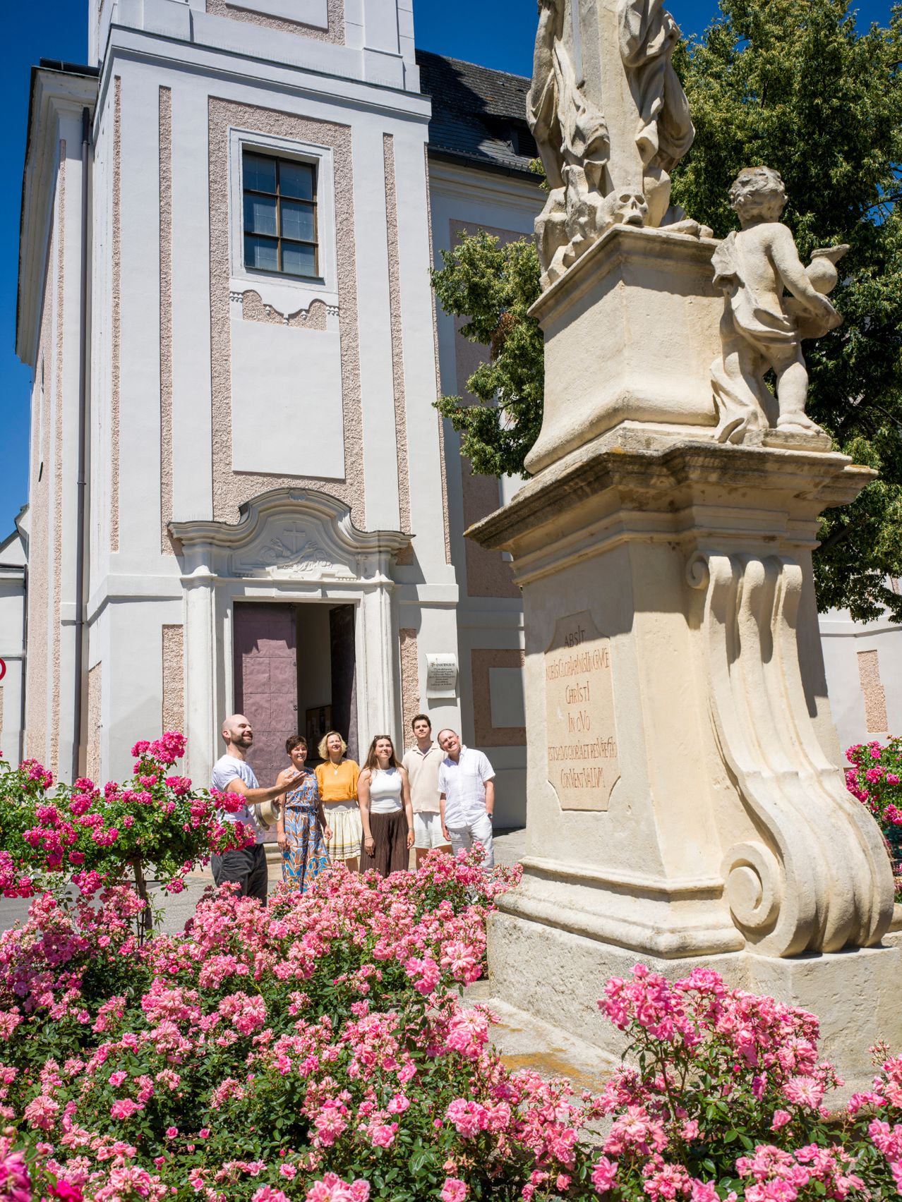 Eine Gruppe von Menschen steht vor einem historischen Gebäude mit einer Statue und rosa Blumen. Das Gebäude hat eine rosa Fassade und ein großes Fenster.