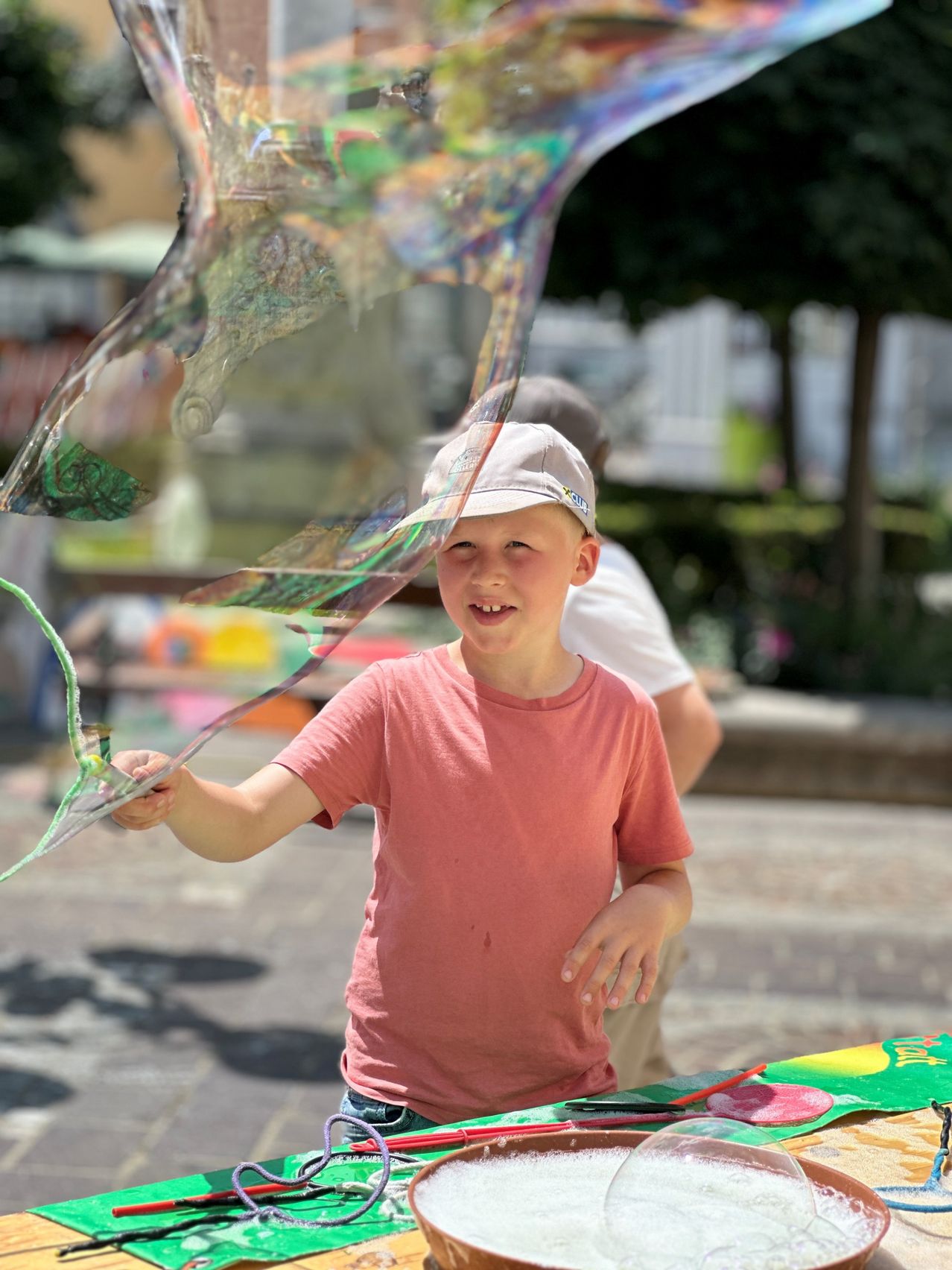 Ein junger Junge in einem rosa T-Shirt und einer Kappe hält eine große Seifenblase im Freien. Bäume und ein unscharfer Hintergrund sind hinter ihm.