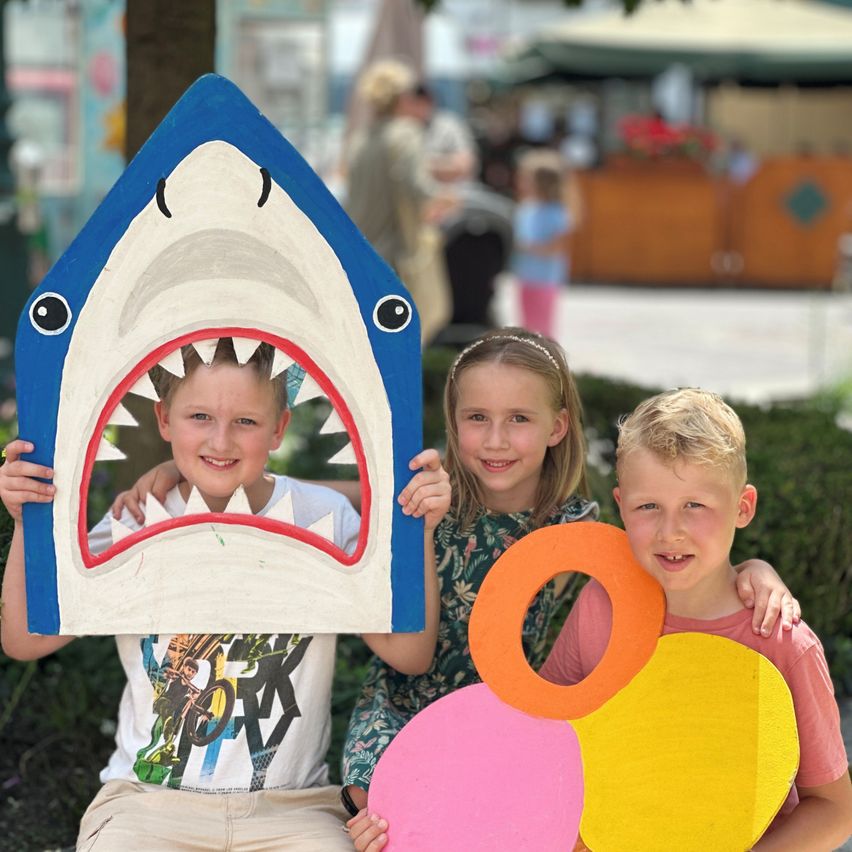 Drei Kinder posieren für ein Foto mit einer Papp-Haimaske und bunten Kreisen und lächeln in die Kamera in einer Außenumgebung.