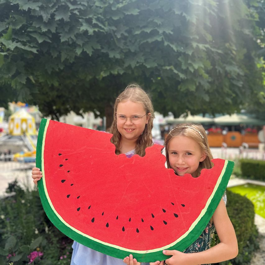 Zwei junge Mädchen halten draußen eine riesige Wassermelonenattrappe, lächeln, mit einem Baum und Pflanzen dahinter.
