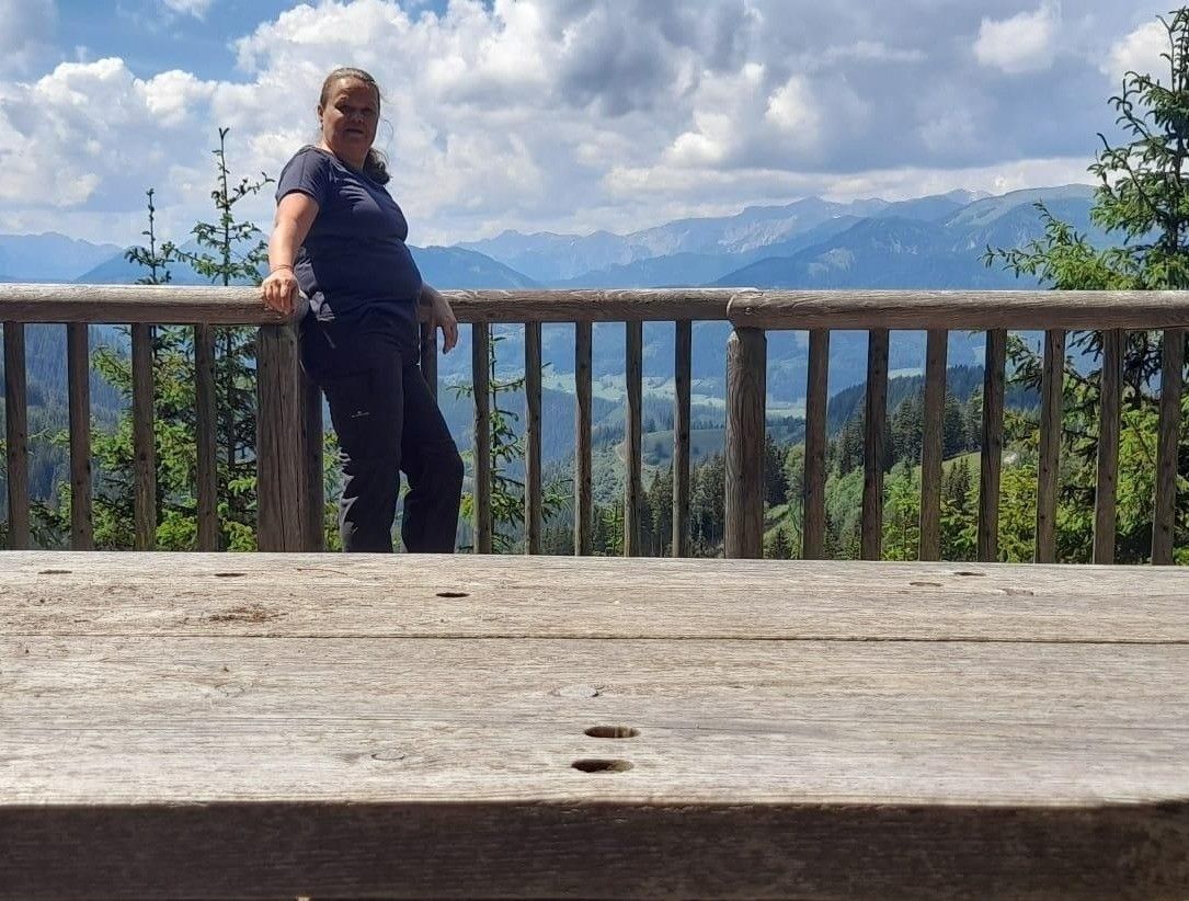 A woman stands on a wooden balcony overlooking a mountainous landscape with a cloudy sky.