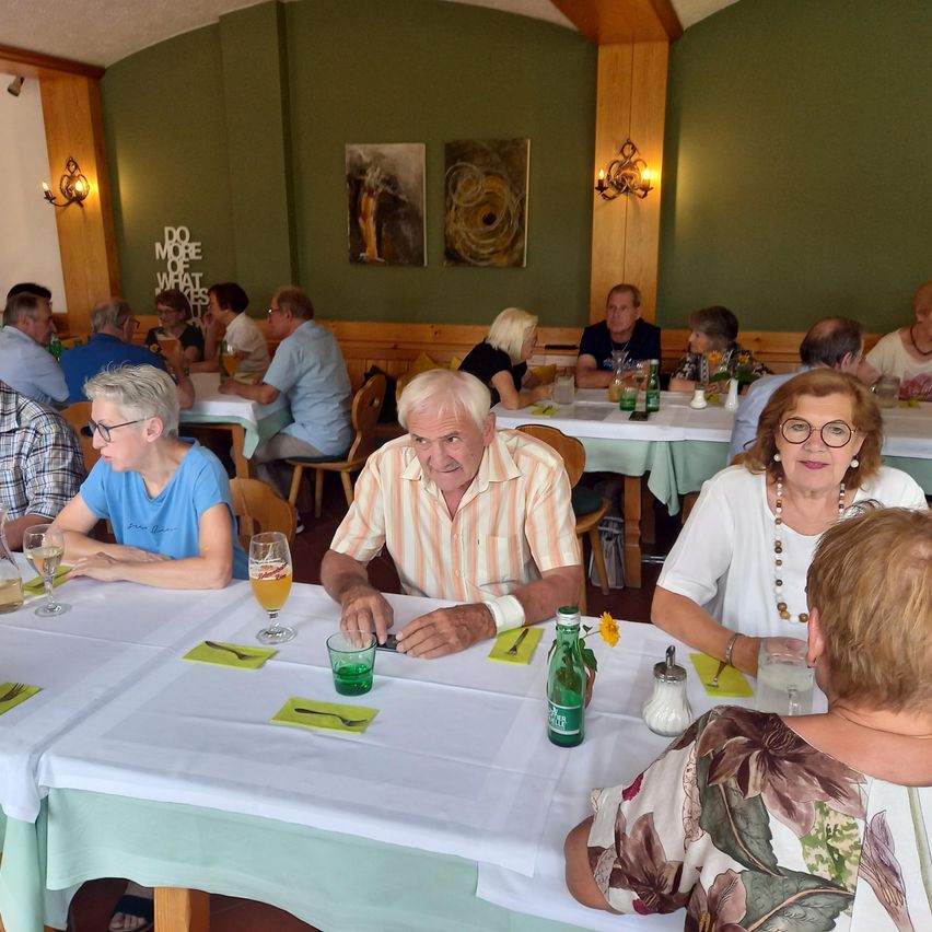 Eine Gruppe von Menschen sitzt in einem Restaurant an Tischen, einige tragen Brillen und Halsketten. Getränke und Teller stehen auf dem Tisch, und Kunst hängt an den Wänden.