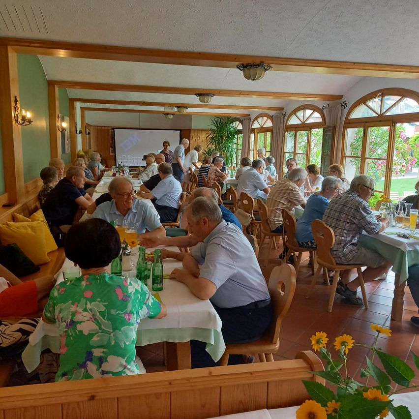 Eine Gruppe von Menschen sitzt an Tischen in einem Restaurant, einige unterhalten sich, während andere essen. Flaschen und Gläser stehen auf dem Tisch. Das Restaurant hat Holzbalken und Glasfenster mit Pflanzen draußen.