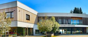 Ein modernes Gebäude mit Glaswänden und Holzfassaden, blauer Himmel darüber und einem großen Baum davor.