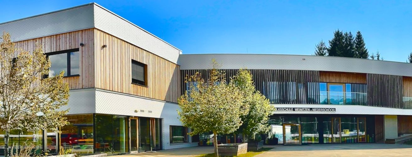 Ein modernes Gebäude mit Glaswänden und Holzfassaden, blauer Himmel darüber und einem großen Baum davor.