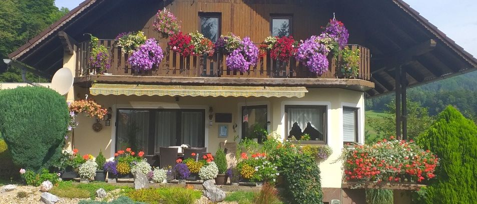 Ein gemütliches Haus mit Balkon und Markise, umgeben von einem Garten voller bunter Blumen und Grünpflanzen.