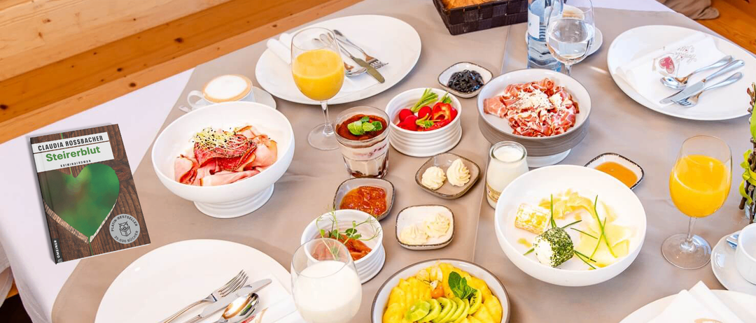 Ein Tisch mit einer Auswahl an Frühstücksgerichten, darunter Obst, Fleisch und Gebäck, mit Gläsern Orangensaft und Milch sowie einem Korb mit Brot.