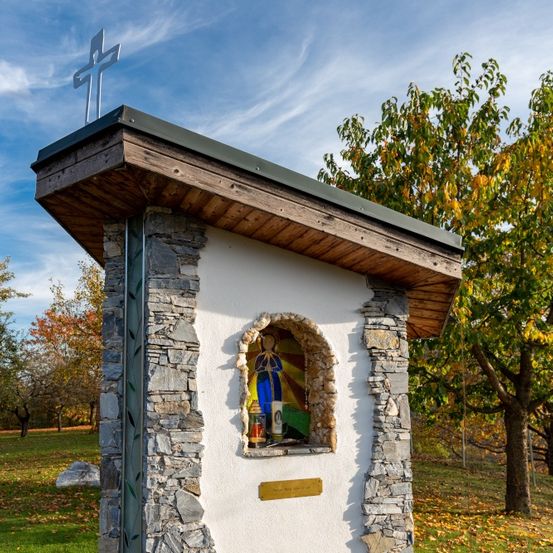Eine kleine Kapelle mit einem Kreuz auf dem Dach steht auf einem Grasfeld. Die Kapelle hat eine Steinmauer und ein Holzdach. Sie hat ein Glasfenster mit einer religiösen Figur im Inneren. Dahinter befinden sich Bäume mit gelben Blättern.