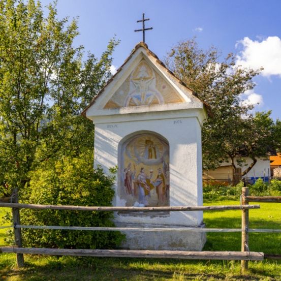 Eine kleine weiße Kapelle mit einem Kreuz auf dem Dach, umgeben von einem Holzzaun und Grün, zeigt ein kunstvolles Gemälde an ihrer Fassade.