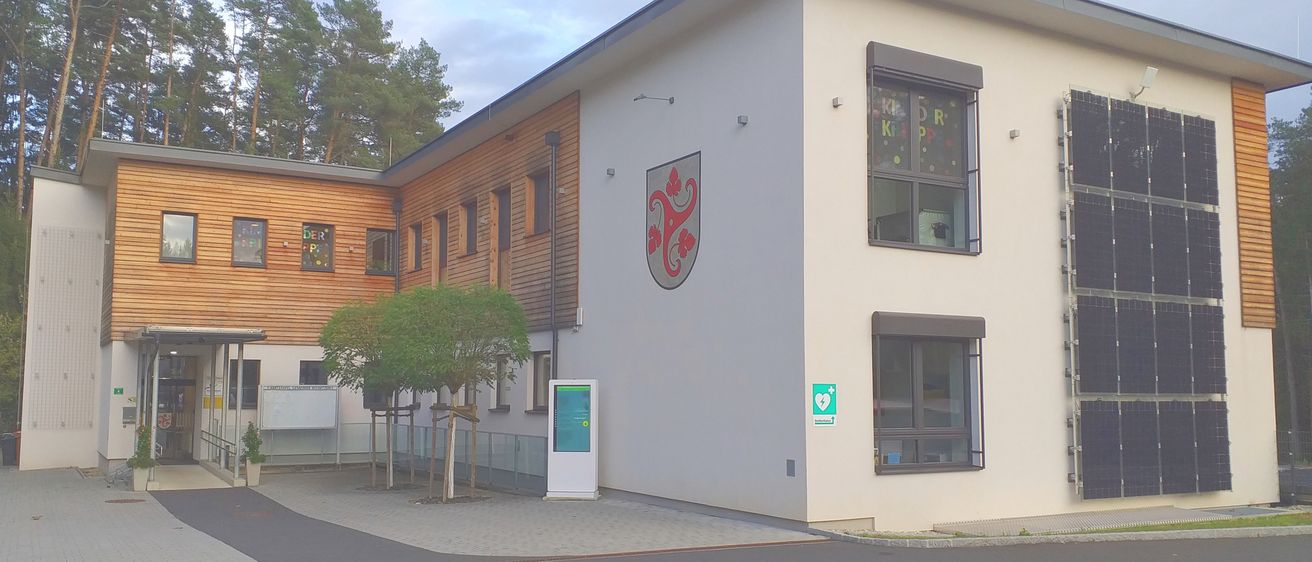 Das Äußere eines modernen Gebäudes mit Logo, Fenstern und einer Tafel. Bäume und ein kleines Infoboard sind davor.