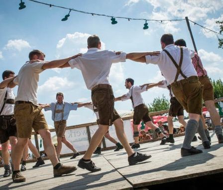 Mehrere Männer in traditioneller bayerischer Kleidung führen einen Tanz auf einer Holzbühne auf, umgeben von Zuschauern und einem bewölkten Himmel.