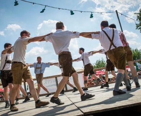 Mehrere Männer in traditioneller bayerischer Kleidung führen einen Tanz auf einer Holzbühne auf, umgeben von Zuschauern und einem bewölkten Himmel.