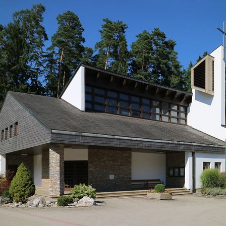 Eine moderne Kirche mit Turm und Kreuz. Sie hat ein graues Dach und Ziegelwände. Es gibt Glasfenster und eine Bank draußen. Bäume sind im Hintergrund.