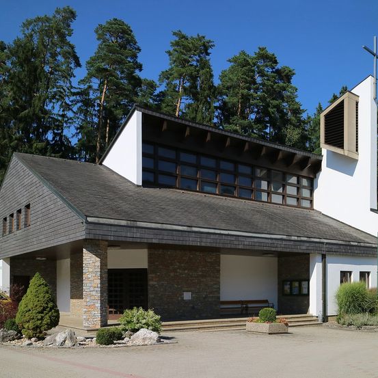 Eine moderne Kirche mit Turm und Kreuz. Sie hat ein graues Dach und Ziegelwände. Es gibt Glasfenster und eine Bank draußen. Bäume sind im Hintergrund.