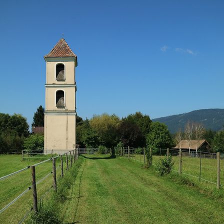 Ein hoher Turm steht in der Mitte eines weiten grünen Feldes, mit Zäunen und Bäumen dahinter. In der Ferne sind ein Haus und ein Berg unter einem klaren blauen Himmel zu sehen.