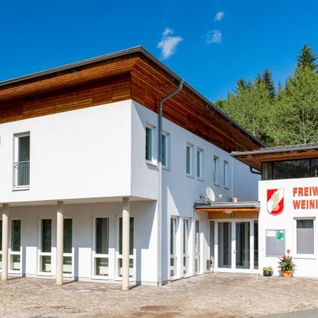 Die Vorderseite eines weißen Gebäudes mit braunem Dach und Fenstern, es steht Freiwillige Feuerwehr Weinitzen-Oberschö in Rot auf der Wand.