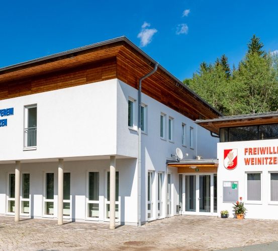 Die Vorderseite eines weißen Gebäudes mit braunem Dach und Fenstern, es steht Freiwillige Feuerwehr Weinitzen-Oberschö in Rot auf der Wand.