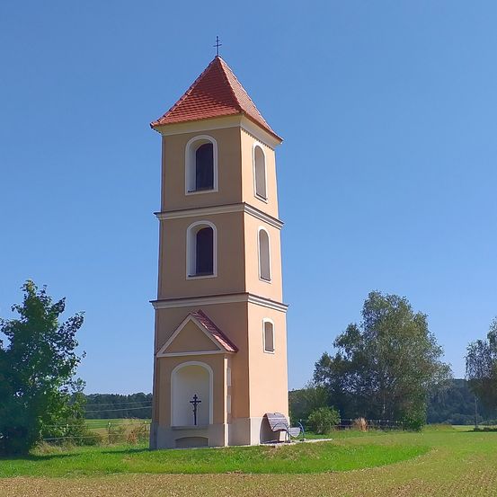 Ein hoher, beigefarbener Turm mit einem roten Dach und Fenstern steht vor einem blauen Himmel. Eine Bank und ein Kreuz stehen davor.